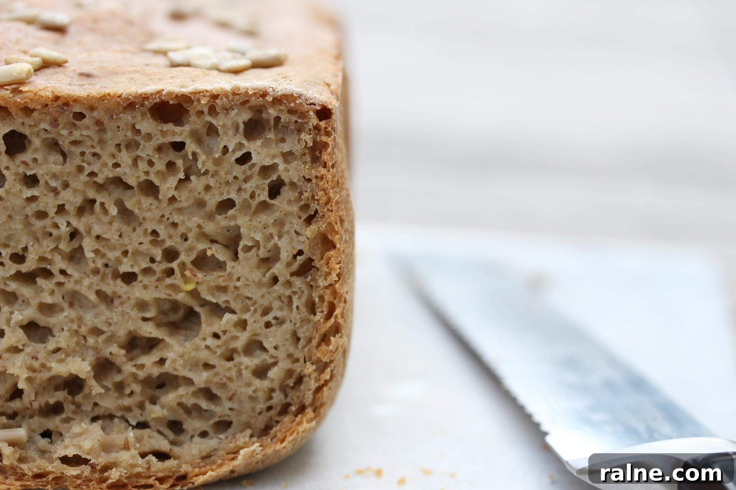 Gluten-Free Vegan Bread Machine Recipe: Soft Loaf Guide 2 A close up of the crumb of the bread. A loaf is to the left of the shot with a knife lying to the right. The crust is cut off the end of the loaf so the texture is in focus and visible.