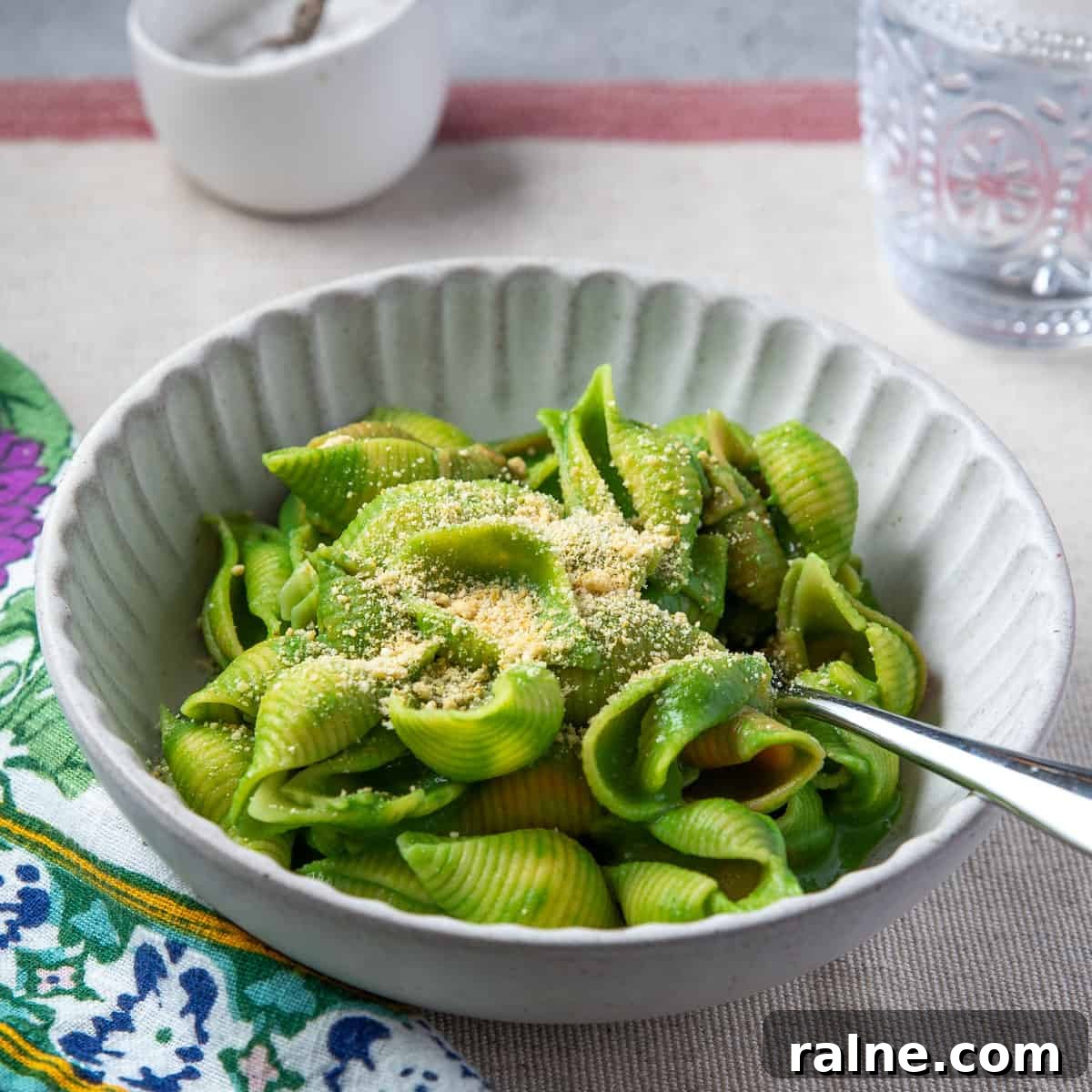 Square photo of vegan spinach pasta in a cream coloured bowl on a table mat with patterned napkin to the side and nutty cheese sprinkled on top.