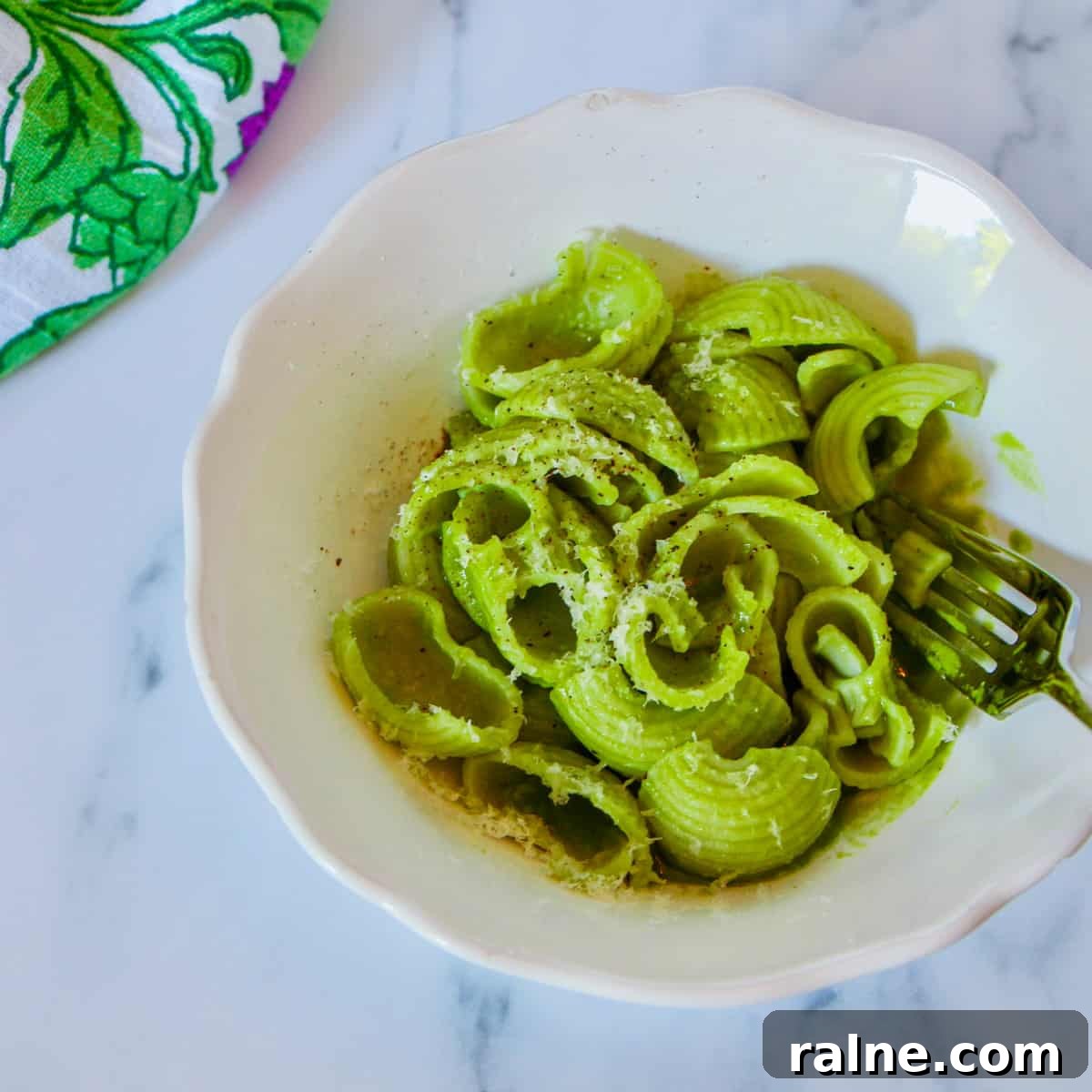 Photo of a white bowl filled with quick Vegan Creamy Spinach Pasta with vegan cheese and pepper sprinkled on top and a patterned napkin in the background, all on a white and grey marble background.