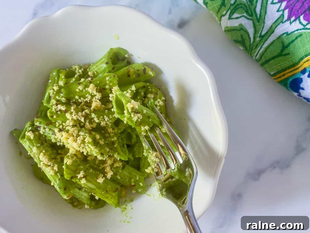photo of a white bowl with creamy spinach pasta and a fork digging in for a mouthful, with nutty cheese and seaweed sprinkled on top and a patterned napkin in the background, all on a white and grey marble countertop.