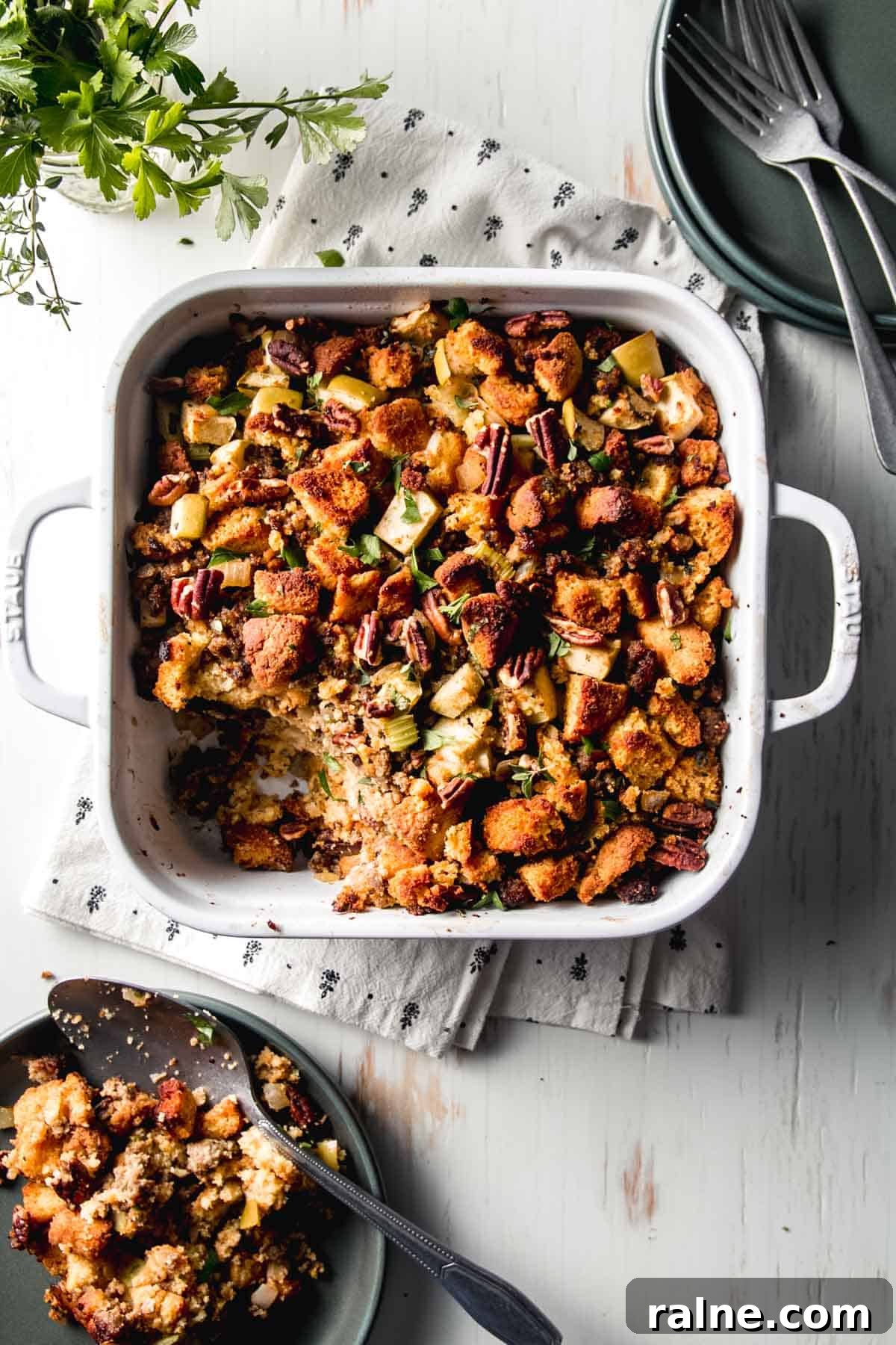 Gluten-Free Cornbread Stuffing with Sausage and Apples for Holiday Meals 2 Gluten-free cornbread stuffing in a white casserole dish next to green plates, fresh herbs, and forks.