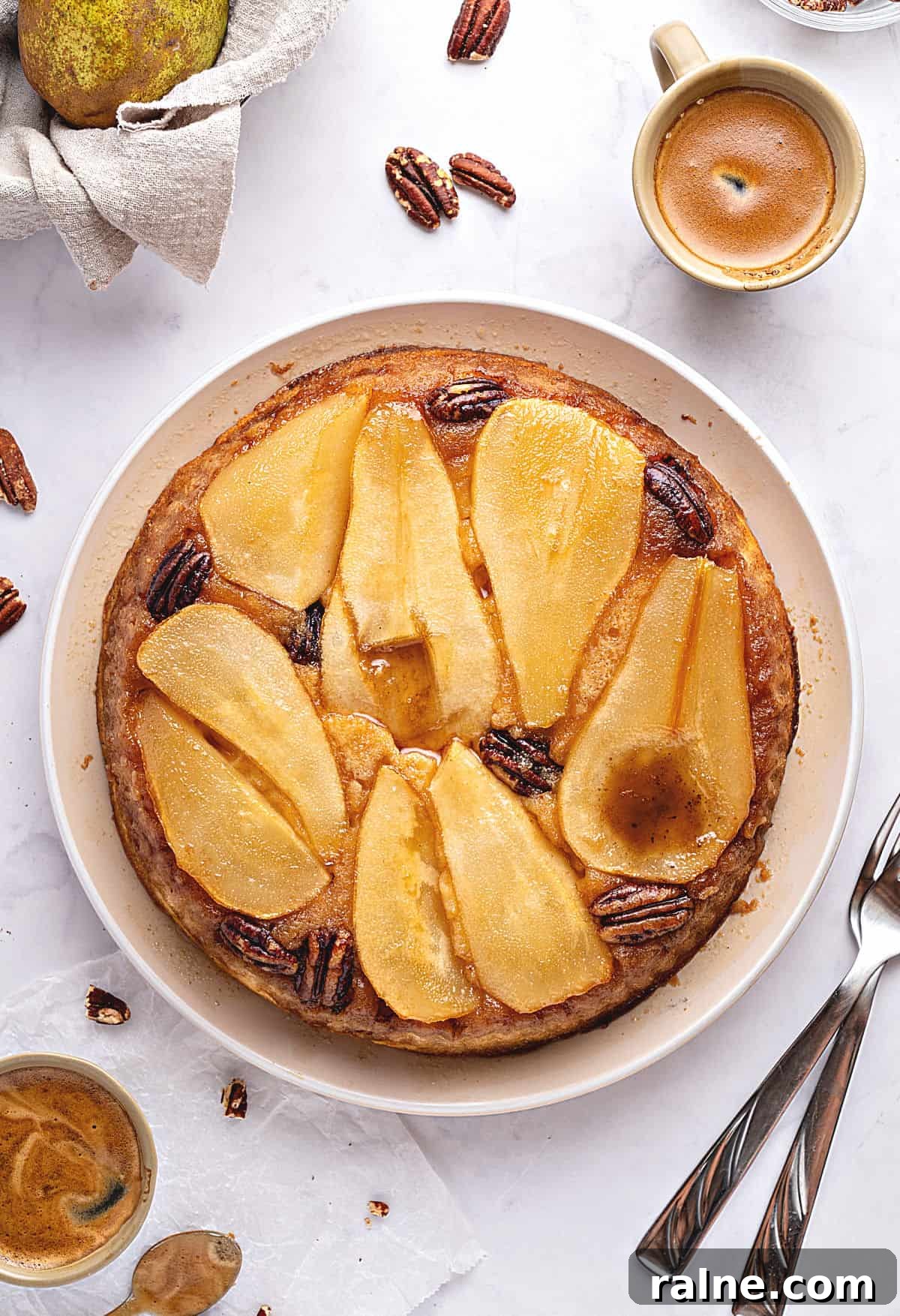 Whole pear upside down cake on a white plate. Top view. Whitish background. Pecans, coffee cups, silver forks.