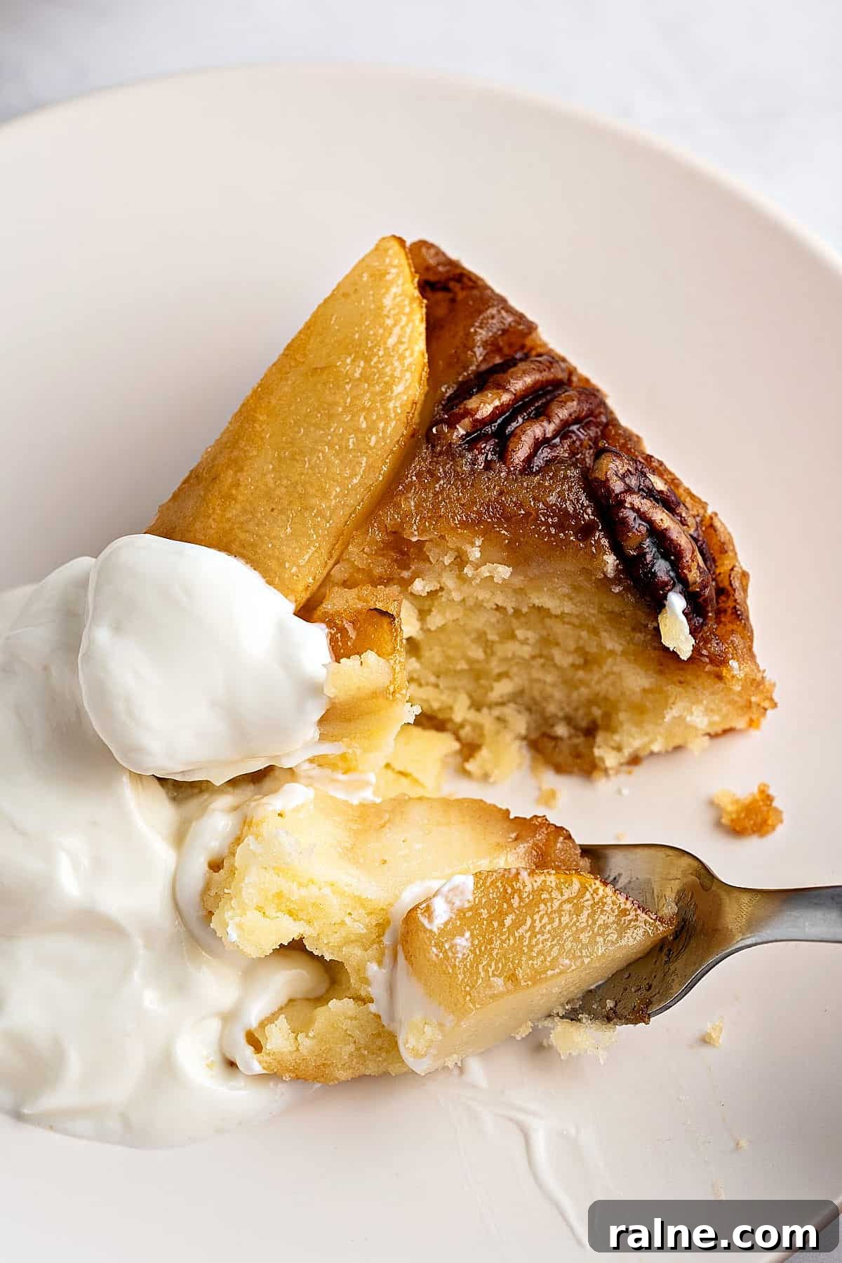 Forking a piece of pear cake with whipped cream on a whitish plate.