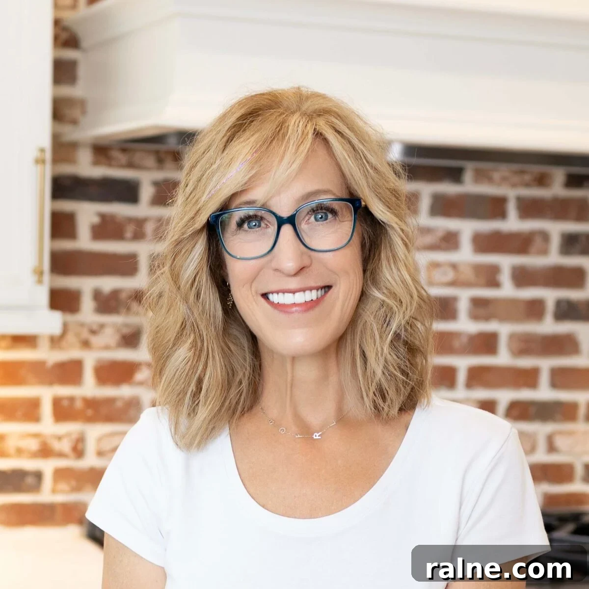 Quick Chicken Dinners for Busy Weeknights 4 Person with shoulder-length blond hair and blue glasses smiling indoors in front of a red brick wall.