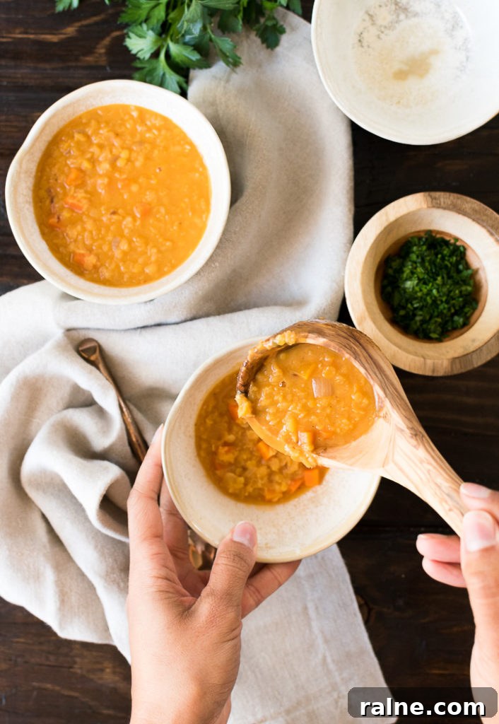 Creamy Coconut Red Lentil Soup with Warm Spices 2 a woman spoons a serving of red lentil soup into a white bowl above a dark wood table with another bowl of soup