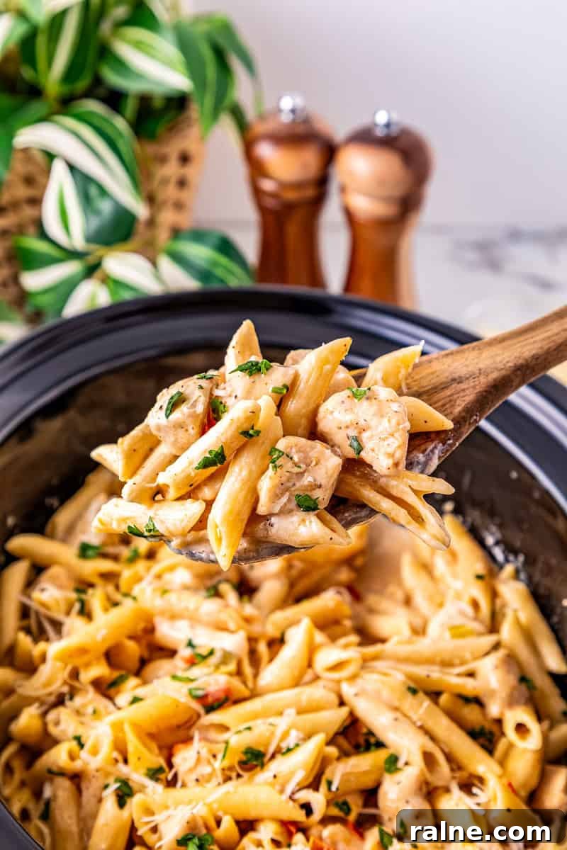 Slow Cooker Cajun Chicken Pasta Recipe for Flavorful Meals 2 a wooden spoon with cajun chicken and penne pasta over top of a black crock pot.