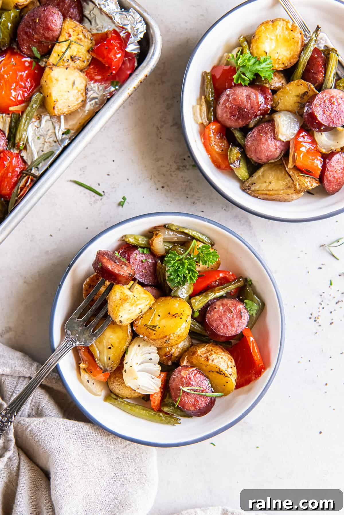 Two bowls filled with roasted sausage and potatoes with green beans and bell peppers.