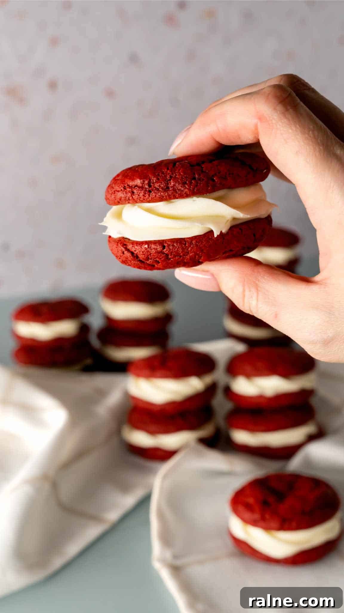 Decadent Red Velvet Whoopie Pies Recipe for Perfect Sandwich Cookies 6 A hand holding up a red velvet whoopie pie with whoopie pies stacked in the background.