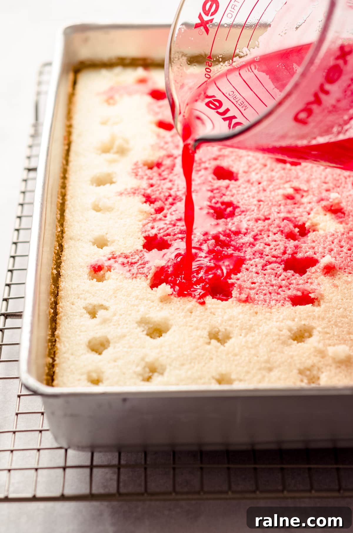 Strawberry Jello Poke Cake Recipe — Moist, Creamy Dessert 11 pouring strawberry jello over sheet cake to make strawberry poke cake