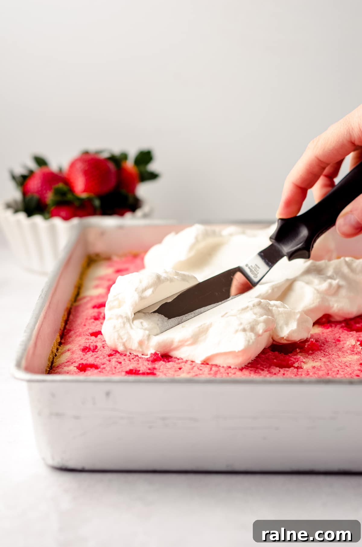 Strawberry Jello Poke Cake Recipe — Moist, Creamy Dessert 14 spreading whipped cream on top of strawberry poke cake in a sheet pan