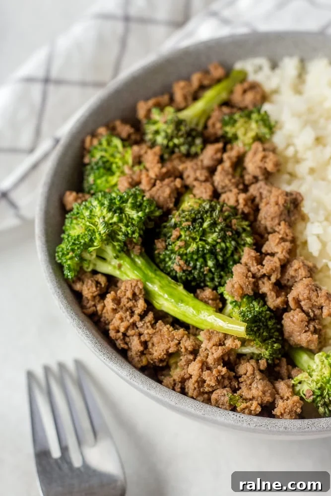 Zoomed in shot of ground beef and broccoli in a gray bowl with cauliflower rice. A silver fork is in the foreground.