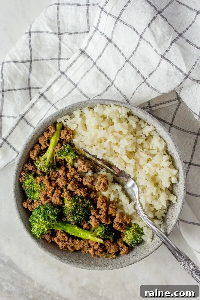 Top down shot of ground beef and broccoli stir fry in gray bowl with cauliflower rice. A fork is in the bowl and a black and white paned cloth napkin is next to the bowl.