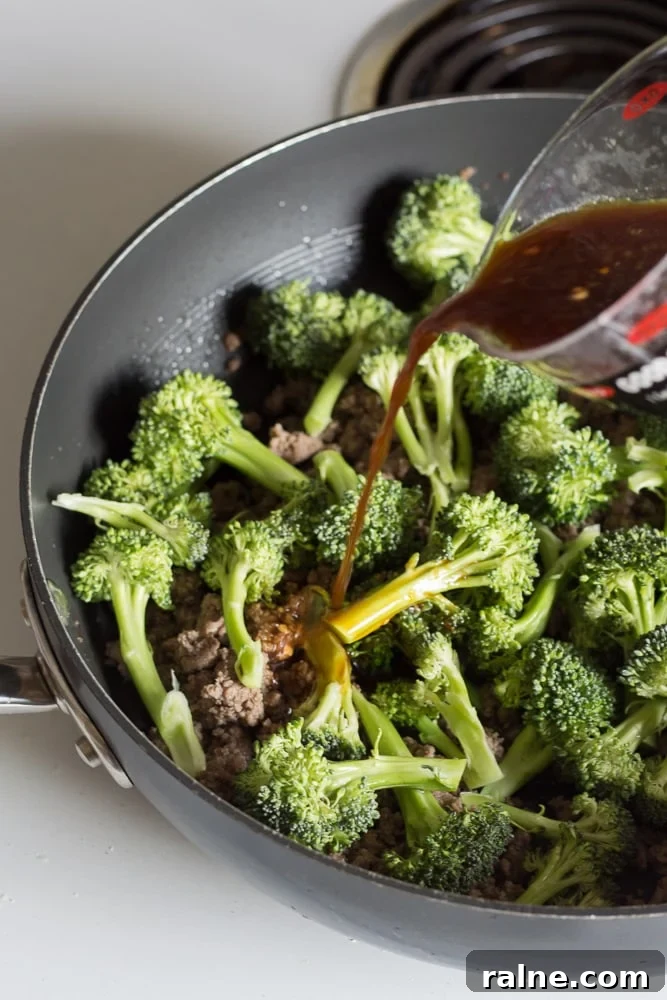 Stir fry sauce being poured over the ground beef and broccoli in a large black pan on the stove.