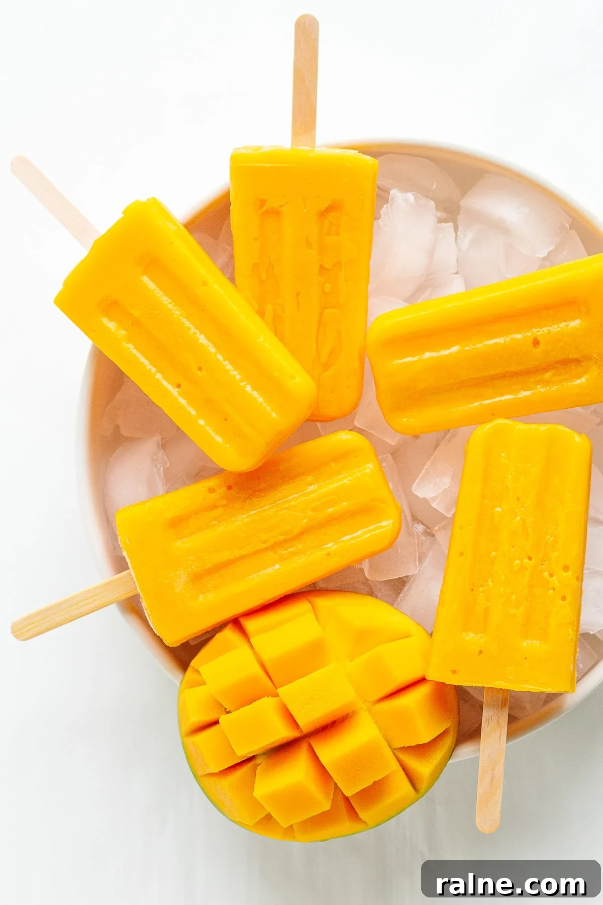 Five mango popsicles and half a fresh mango sitting on a bowl of ice.