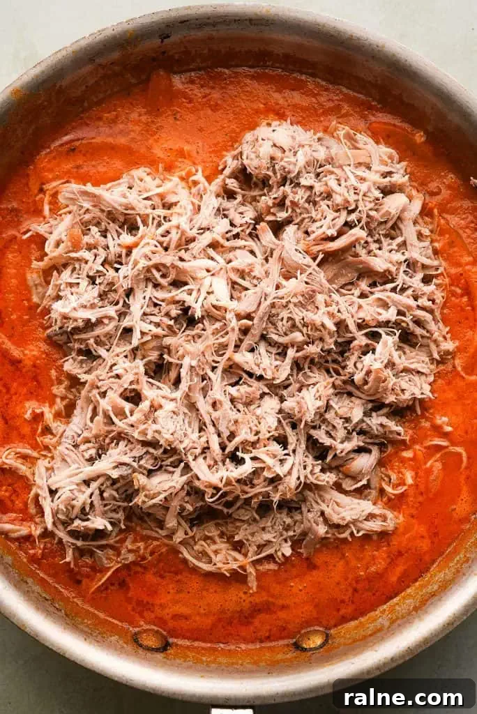 Shredded chicken being added to a skillet of red tinga sauce before being mixed together.