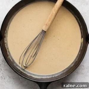 A cast iron skillet filled with a light brown sauce, with a whisk resting inside.