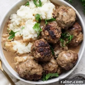A plate of meatballs covered in gravy, served with mashed potatoes and garnished with fresh parsley. A fork and a small bowl of pepper sit adjacent to the plate.