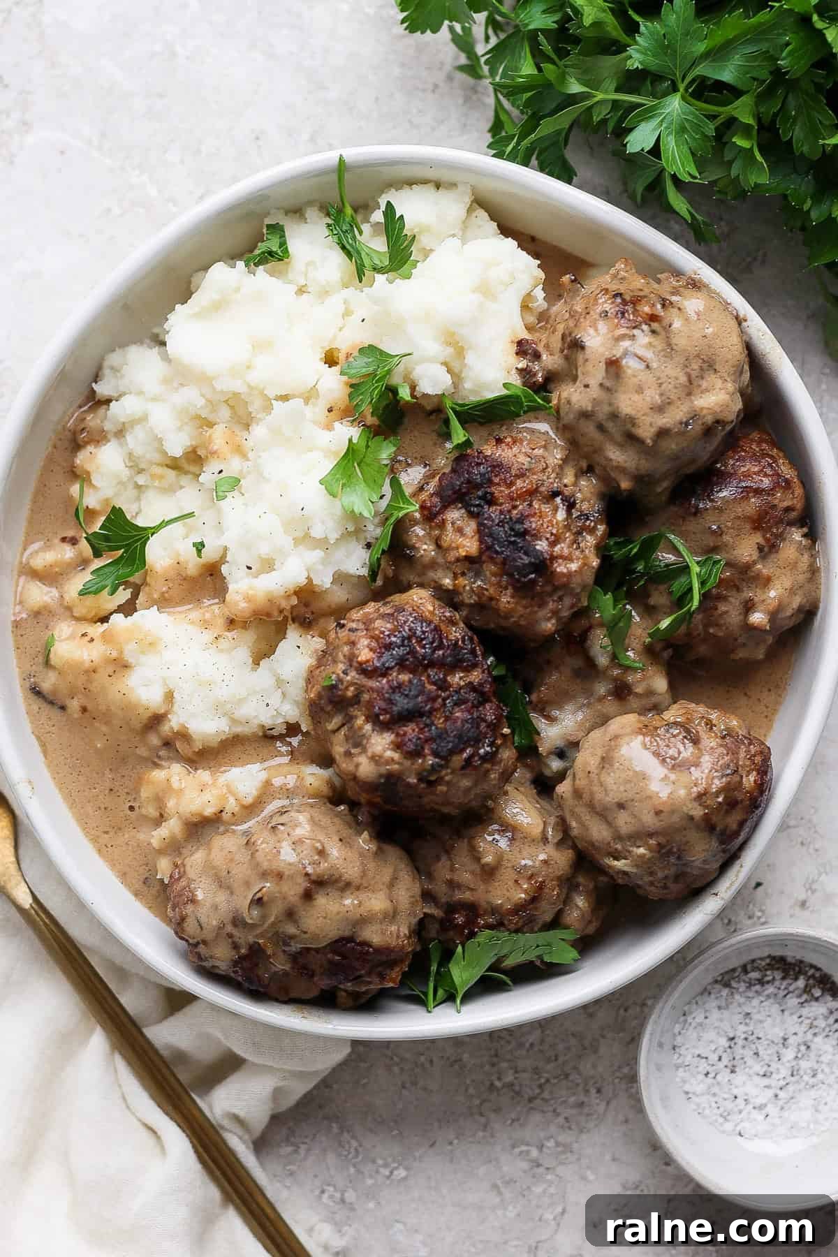 A plate of meatballs covered in gravy, served with mashed potatoes and garnished with fresh parsley. A fork and a small bowl of pepper sit adjacent to the plate.