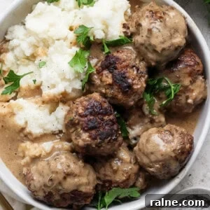 A bowl of meatballs in gravy, served with mashed potatoes, garnished with sprigs of fresh parsley.