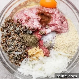 A glass bowl containing ground meat, an egg yolk, wild rice, spices, breadcrumbs, minced garlic, and shredded onions.
