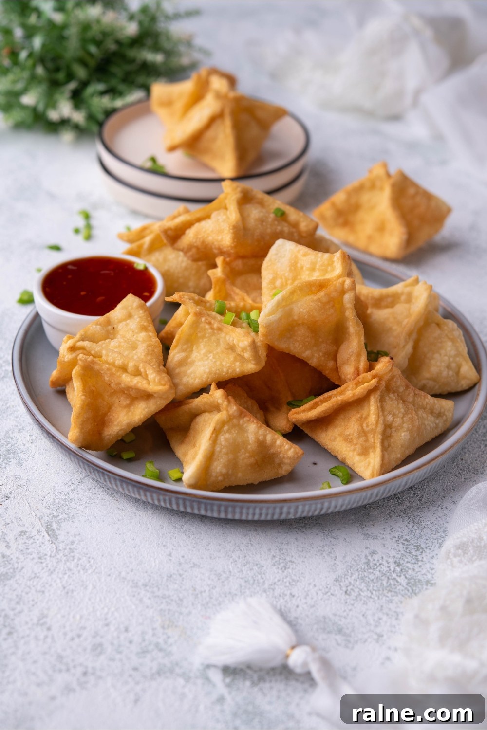 Crispy Cream Cheese Rangoon Wontons: Easy Homemade Recipe 2 A grey plate with Panda Express cream cheese rangoon piled on top of each other and a small bowl of red chili sauce. In the background is a smaller plate of rangoon.
