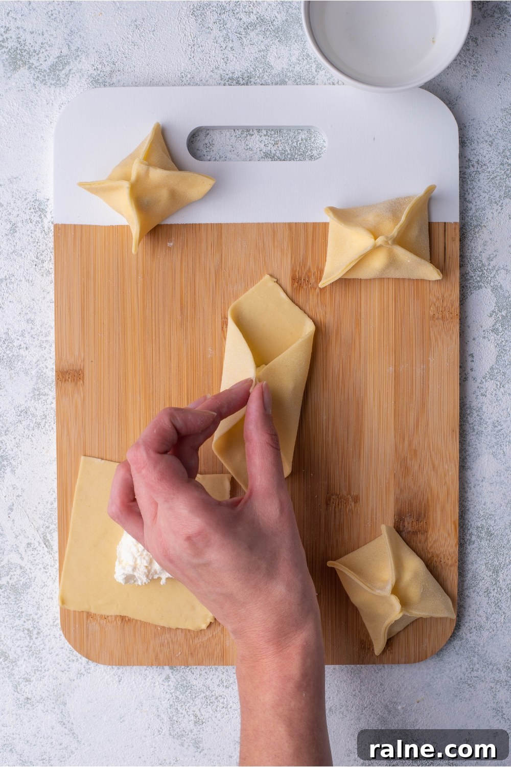 Crispy Cream Cheese Rangoon Wontons: Easy Homemade Recipe 7 Hand pinching two corners of a wonton wrapper that is on a cutting board alongside cream cheese filled rangoon.