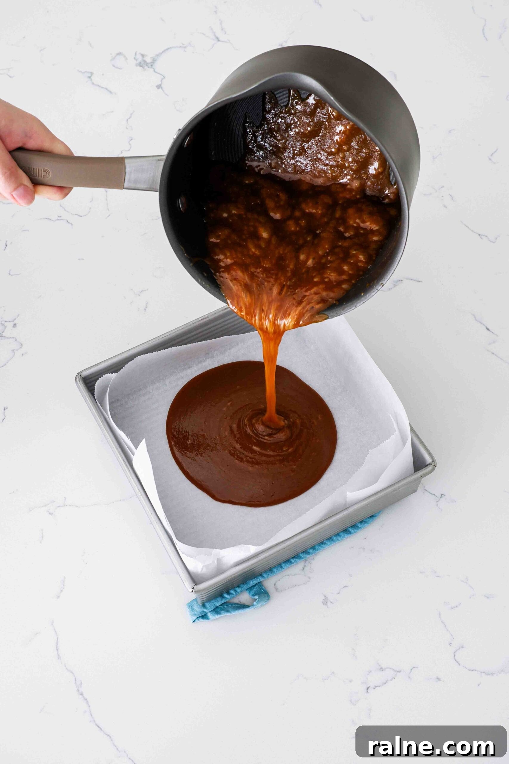 Pumpkin Spice Caramels Recipe: Homemade Fall Candy Guide 11 A hand pours pumpkin spice caramel into a parchment-lined pan.