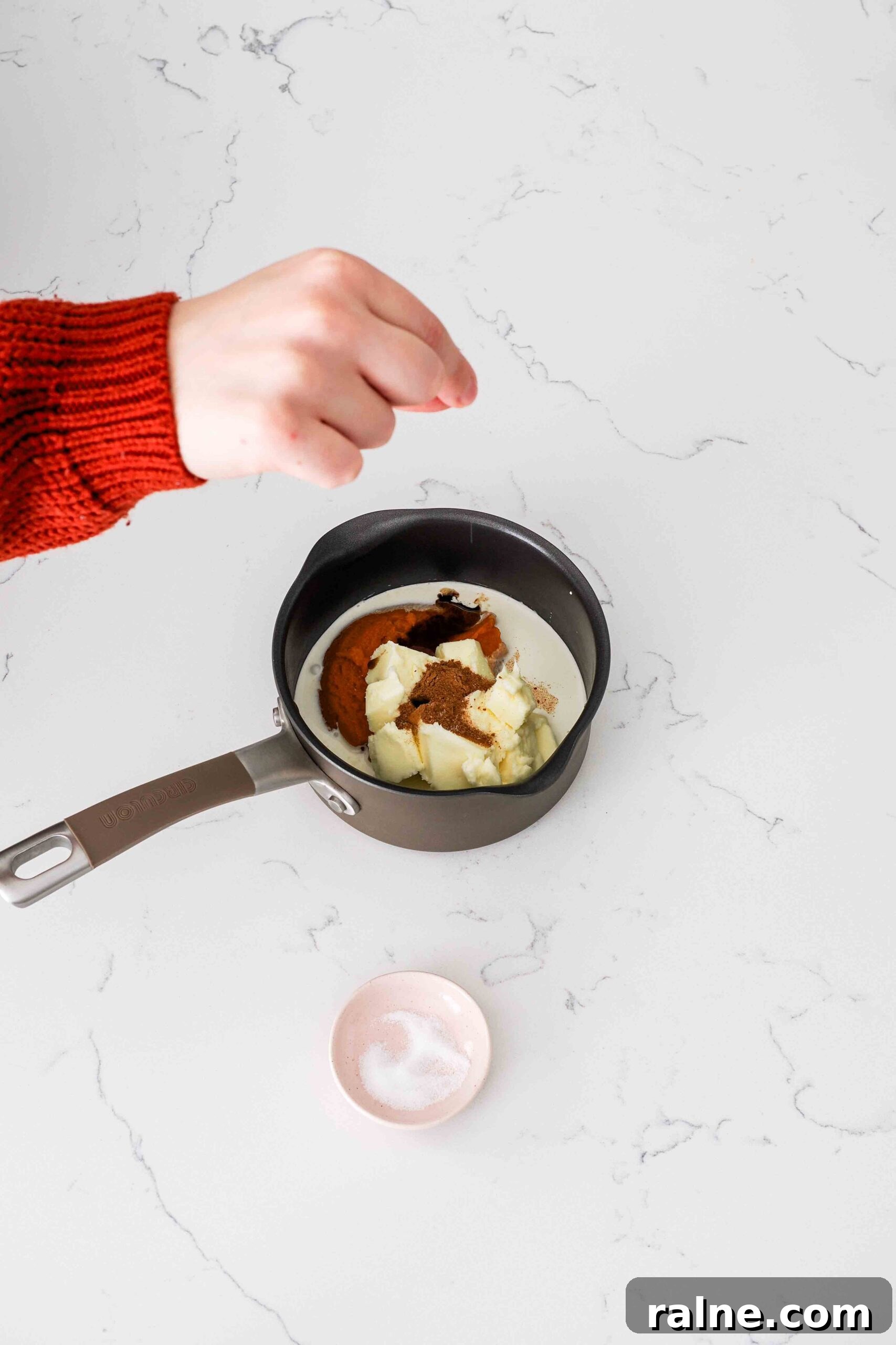 Pumpkin Spice Caramels Recipe: Homemade Fall Candy Guide 5 A hand adds a pinch of salt to a small saucepan with pumpkin, butter, and other ingredients.