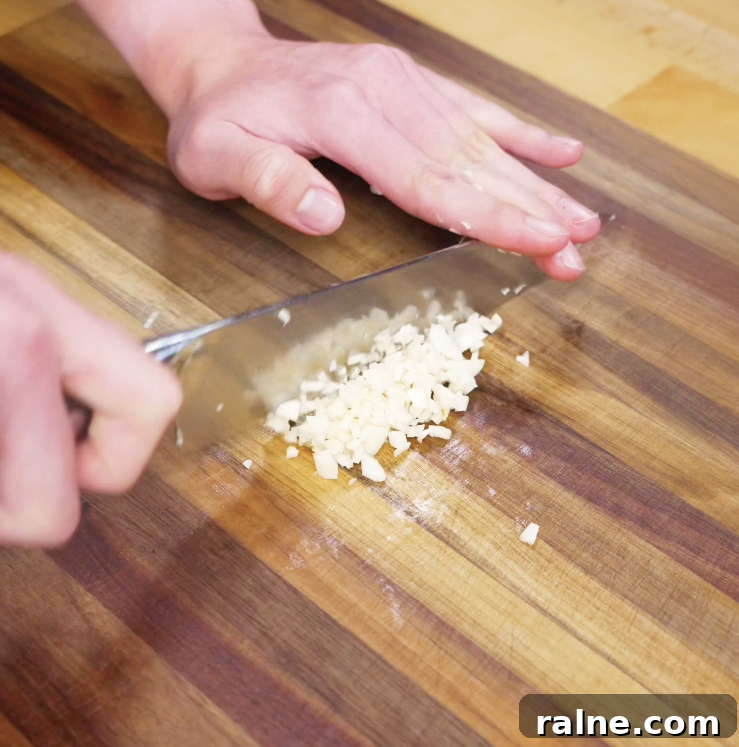 A close-up of hands finely mincing fresh garlic cloves on a wooden cutting board with a sharp knife.