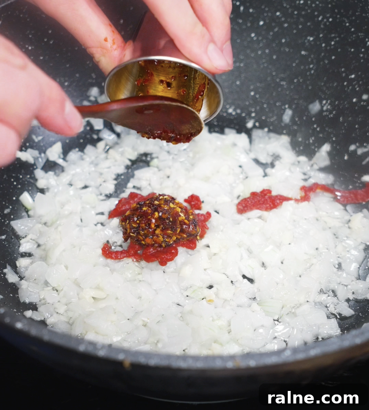 Tomato paste and chili oil being added to the pan of onions and garlic, creating a rich flavor base.