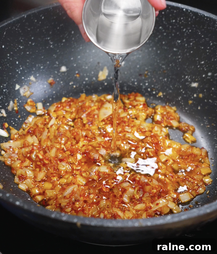 A hand pours clear vodka into the sautéed onion and chili paste mixture to deglaze and add depth.
