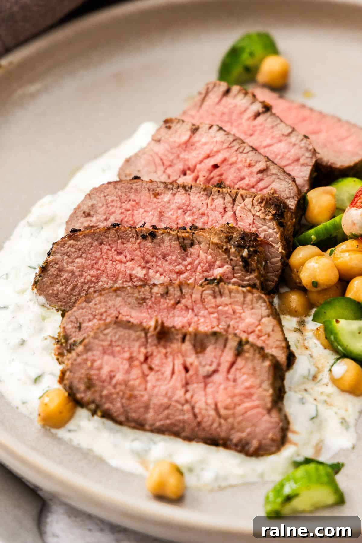 Marinated Lamb Backstrap Recipe: Juicy Grilled Lamb Backstrap 2 Round grey plate with a serving of sliced Lamb Backstrap, sitting on yoghurt sauce, with a chickpea cucumber side salad.