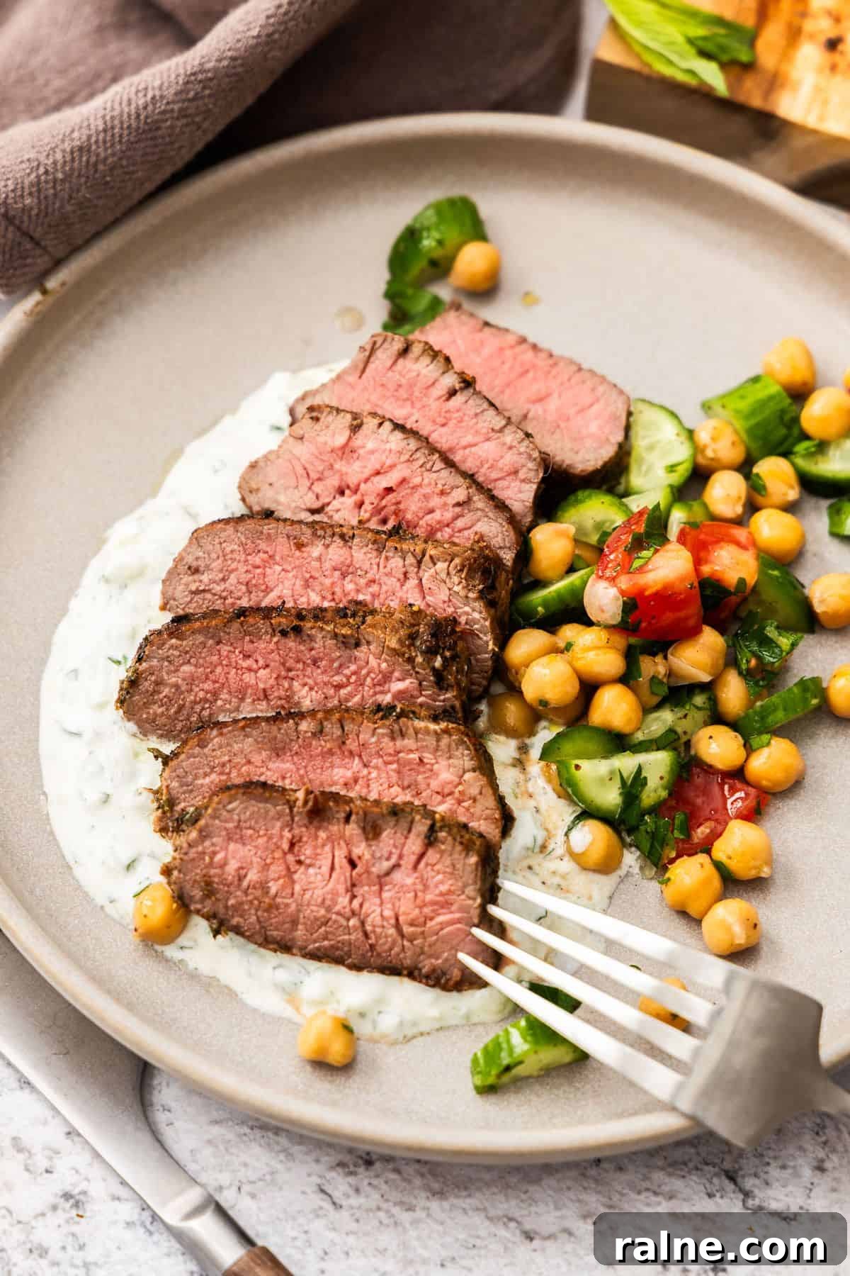 Marinated Lamb Backstrap Recipe: Juicy Grilled Lamb Backstrap 7 Round grey plate with a serving of sliced Lamb Backstrap, sitting on yoghurt sauce, with a chickpea cucumber side salad.