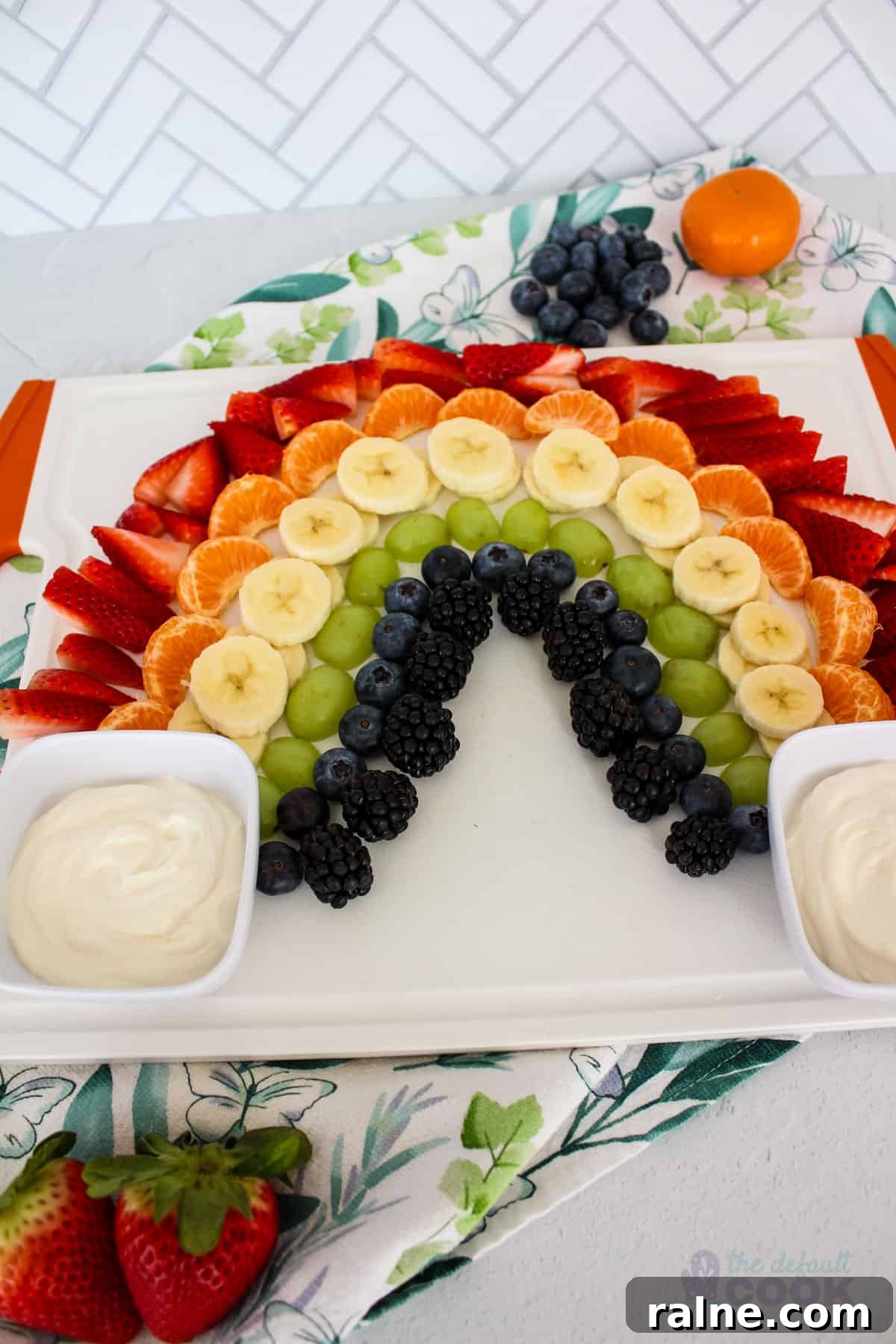 Colorful Fruit Platter with Creamy Yogurt Dip 2 A fruit platter arranged in a rainbow pattern on a white serving tray, with yogurt dips on either side, against a white tile background with a decorative napkin, and 'the default cook' watermark at the bottom right.