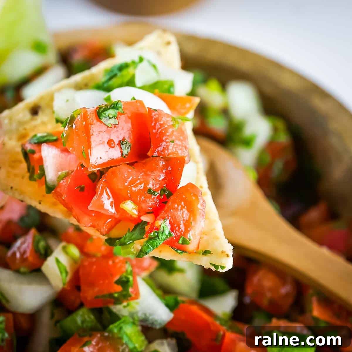 Authentic Mexican Pico de Gallo Recipe: Fresh Salsa Tradicional 5 Side close up shot of a tortilla chip holding a scoop of pico de gallo over a wooden bowl of it with a wooden spoon.