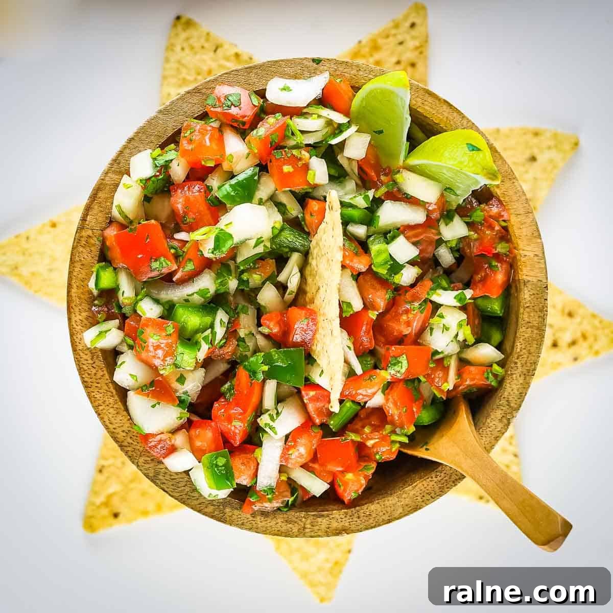 Authentic Mexican Pico de Gallo Recipe: Fresh Salsa Tradicional 6 Overhead shot of a tortilla chip, lime wedges, and a wooden spoon sticking out of a bowl of pico de gallo in a wooden bowl with more tortilla chips arranged in a circle around the bowl on a white surface.