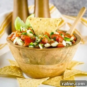 Authentic Mexican Pico de Gallo Recipe: Fresh Salsa Tradicional 7 Side shot of a tortilla chip, lime wedges, and a wooden spoon sticking out of a bowl of pico de gallo in a wooden bowl with more tortilla chips scattered around on a white surface.
