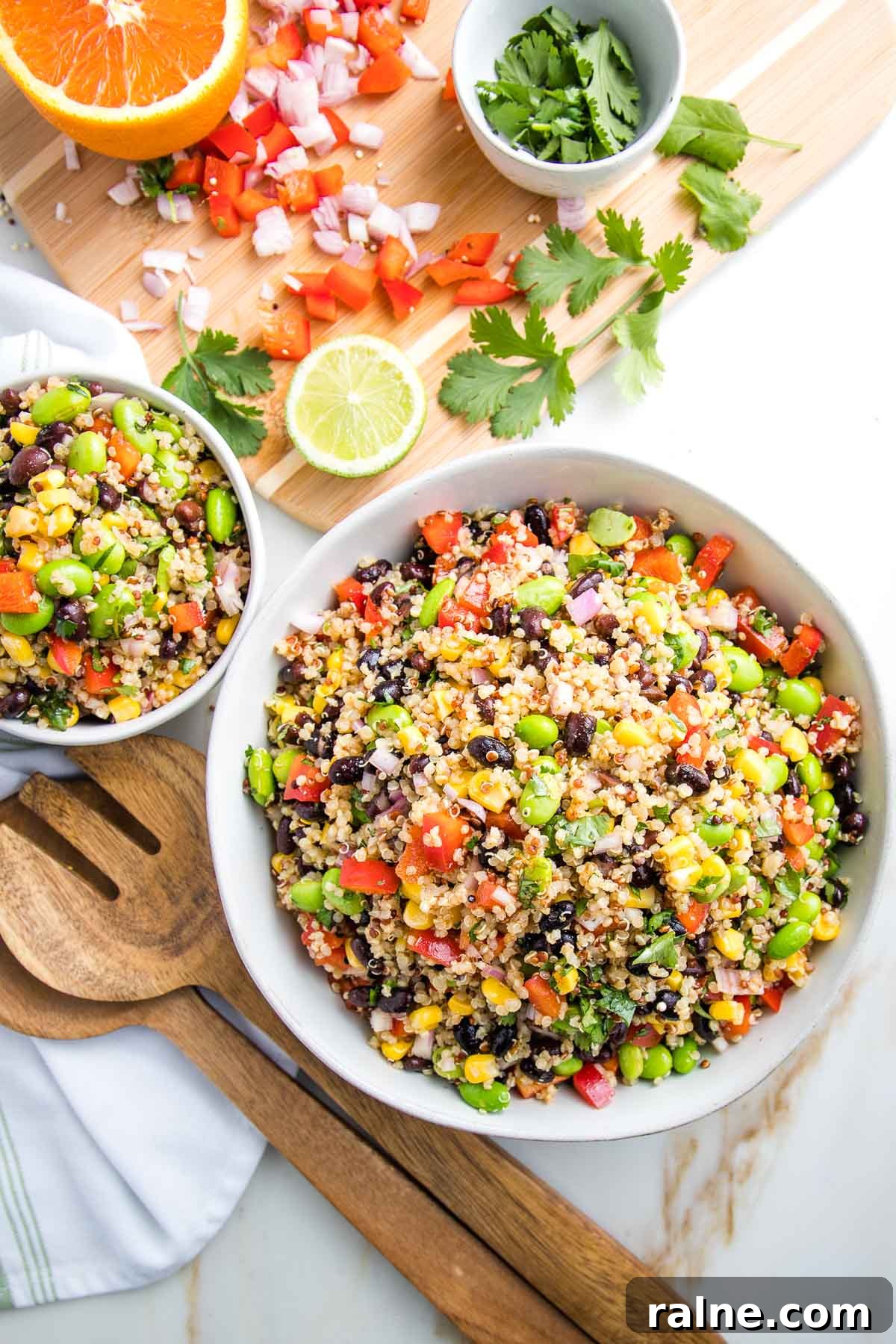Southwest Quinoa Salad with Black Beans, Corn & Lime Dressing 2 Large and small white bowls filled with colorful quinoa salad. In the background there is a cutting board with scraps of chopped red pepper, shallot and fresh cilantro. Also there is half an orange and half a lime. Wooden salad servers are next to the bowls.