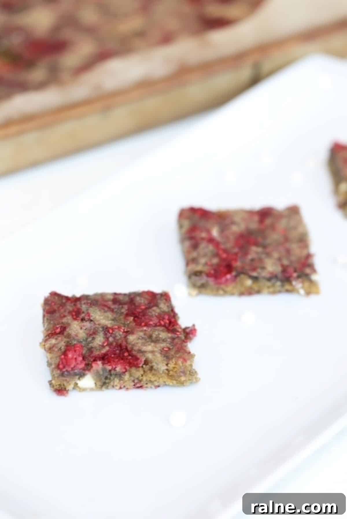 Raspberry White Chocolate Blondies with Crisp Edges and Gooey Center 2 Two raspberry white chocolate blondies on a serving platter with a lined bar pan of blondies in the background.