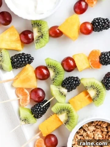 Pieces of fresh fruit on toothpicks and bamboo skewers on a white plate with yogurt and granola in separate ramekins.
