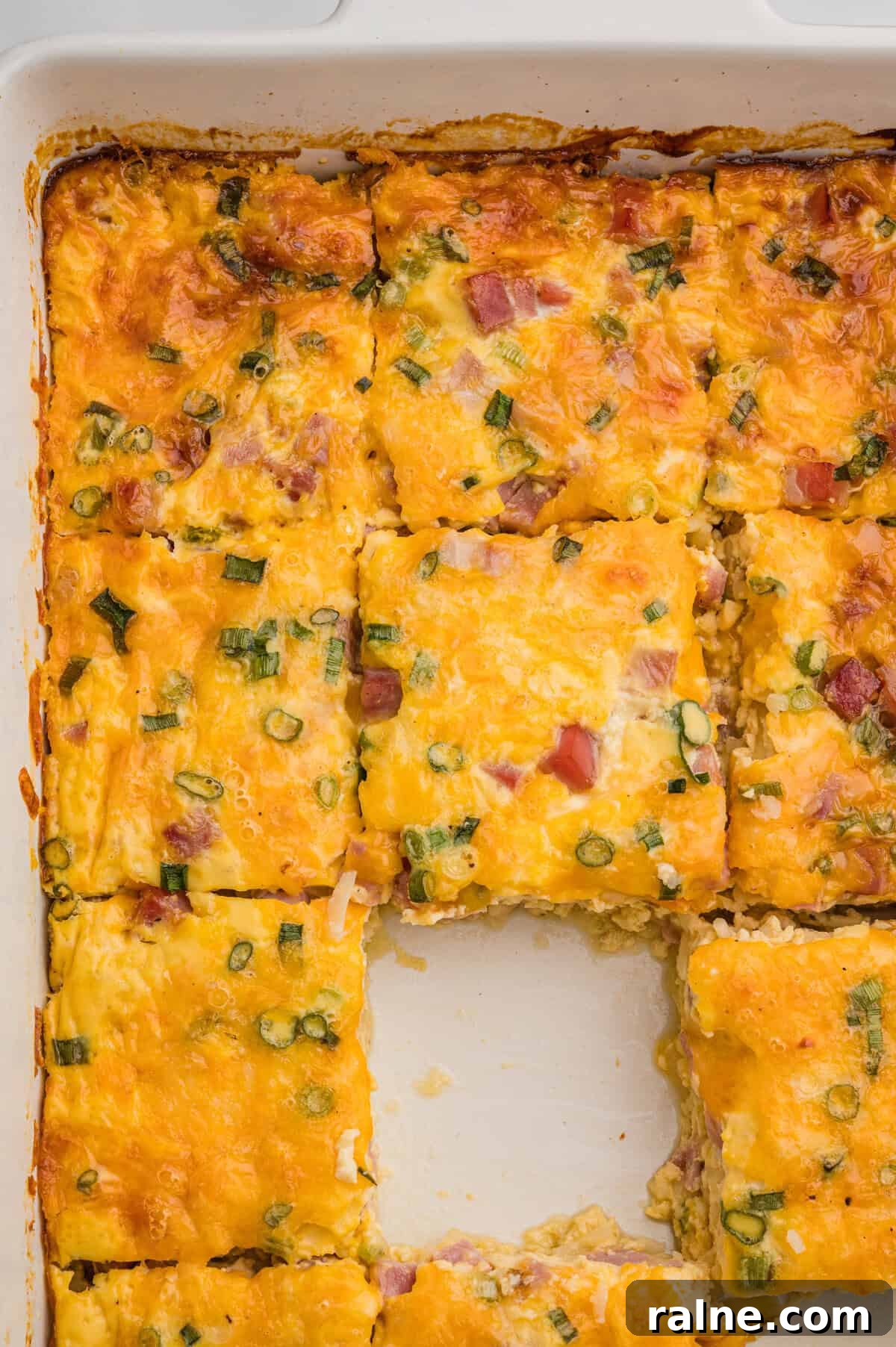 Overhead view of a white baking dish with a sliced breakfast casserole, featuring melted cheese, ham, and green onions, with the center piece removed.