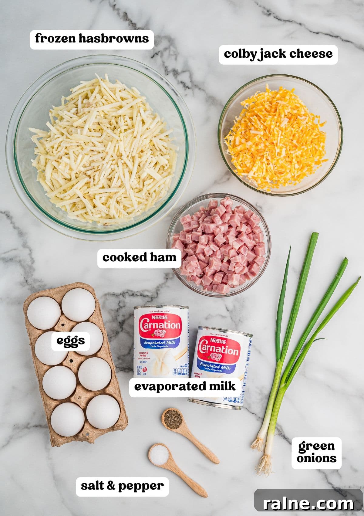 Labeled ingredients on a marble surface, including a bowl of frozen hash browns, shredded Colby Jack cheese, and diced cooked ham, plus eggs, evaporated milk, green onions, and small wooden spoons with salt and pepper.
