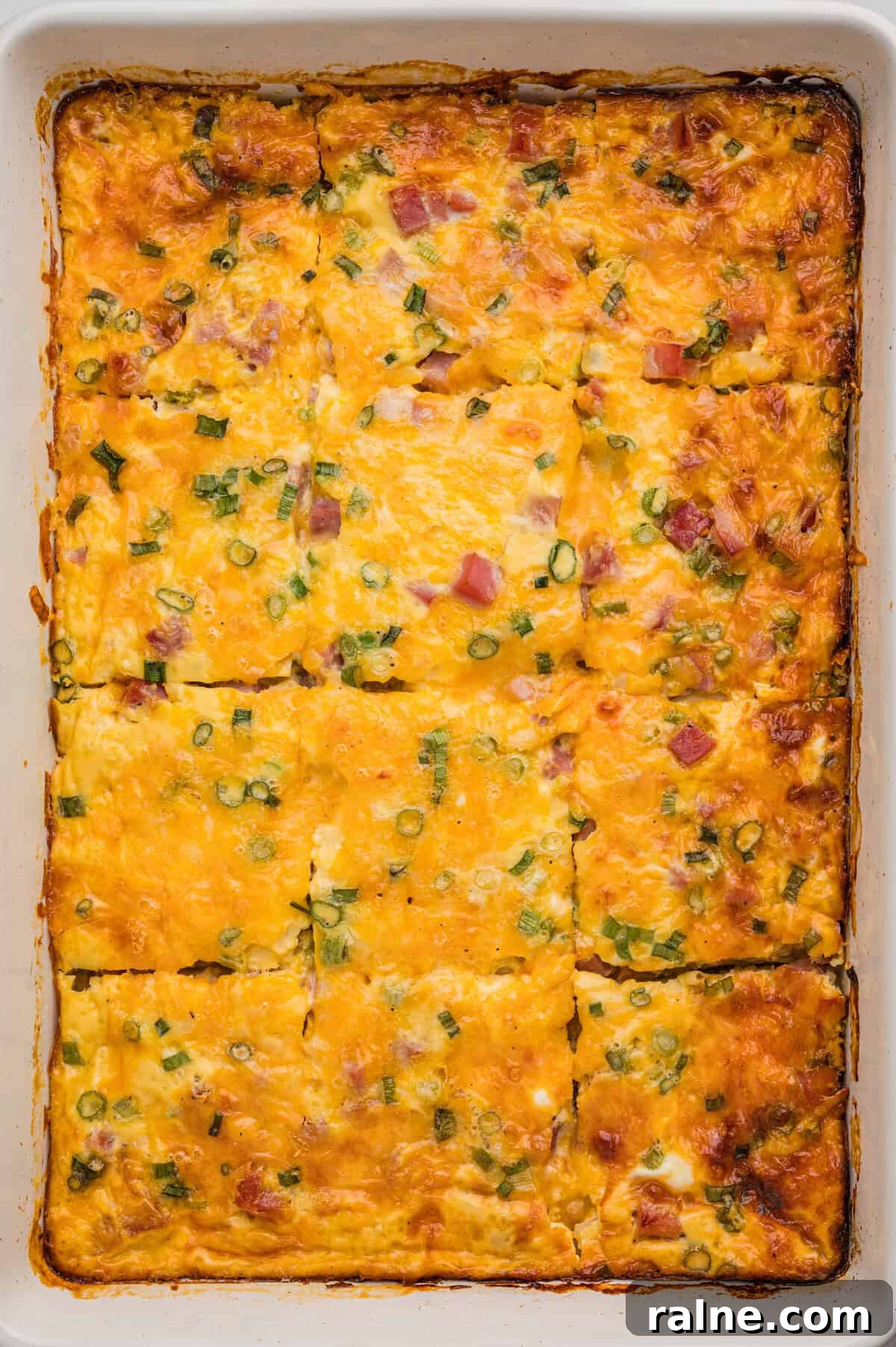A fully baked farmer's casserole in a white dish, cut into rectangular slices with golden melted cheese, diced ham, and green onion visible on top.