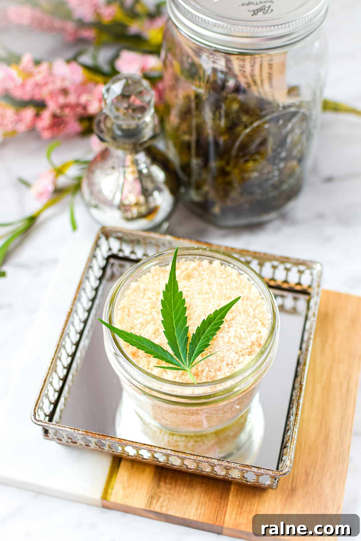 A white counter top with a cutting board with a mirrored tray topped with a mason jar full of cannabis salt, garnished with a cannabis leaf