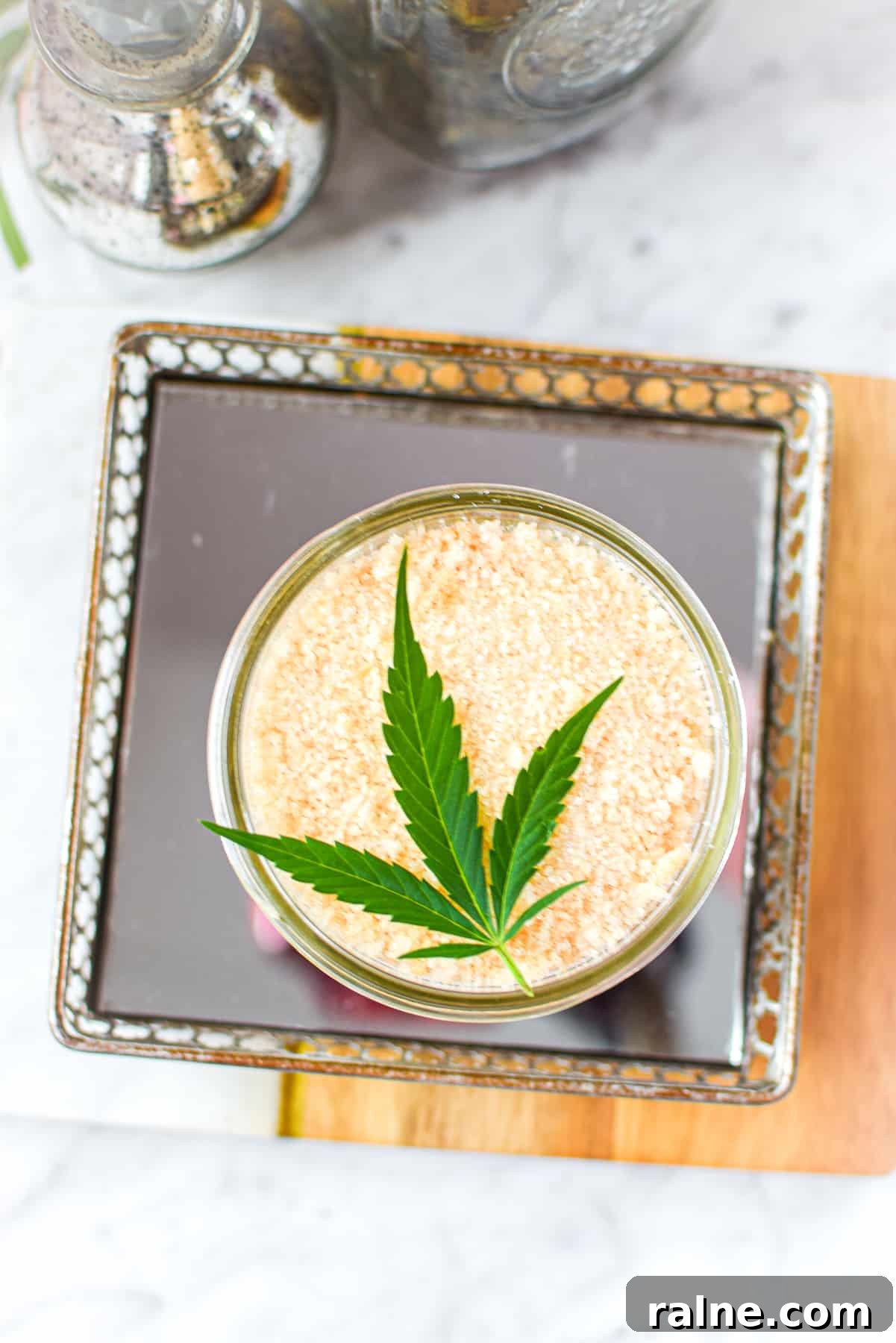 A white counter top with a cutting board with a mirrored tray topped with a mason jar full of cannabis salt, garnished with a cannabis leaf