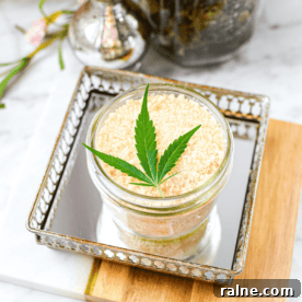 A white counter top with a cutting board with a mirrored tray topped with a mason jar full of cannabis salt, garnished with a cannabis leaf