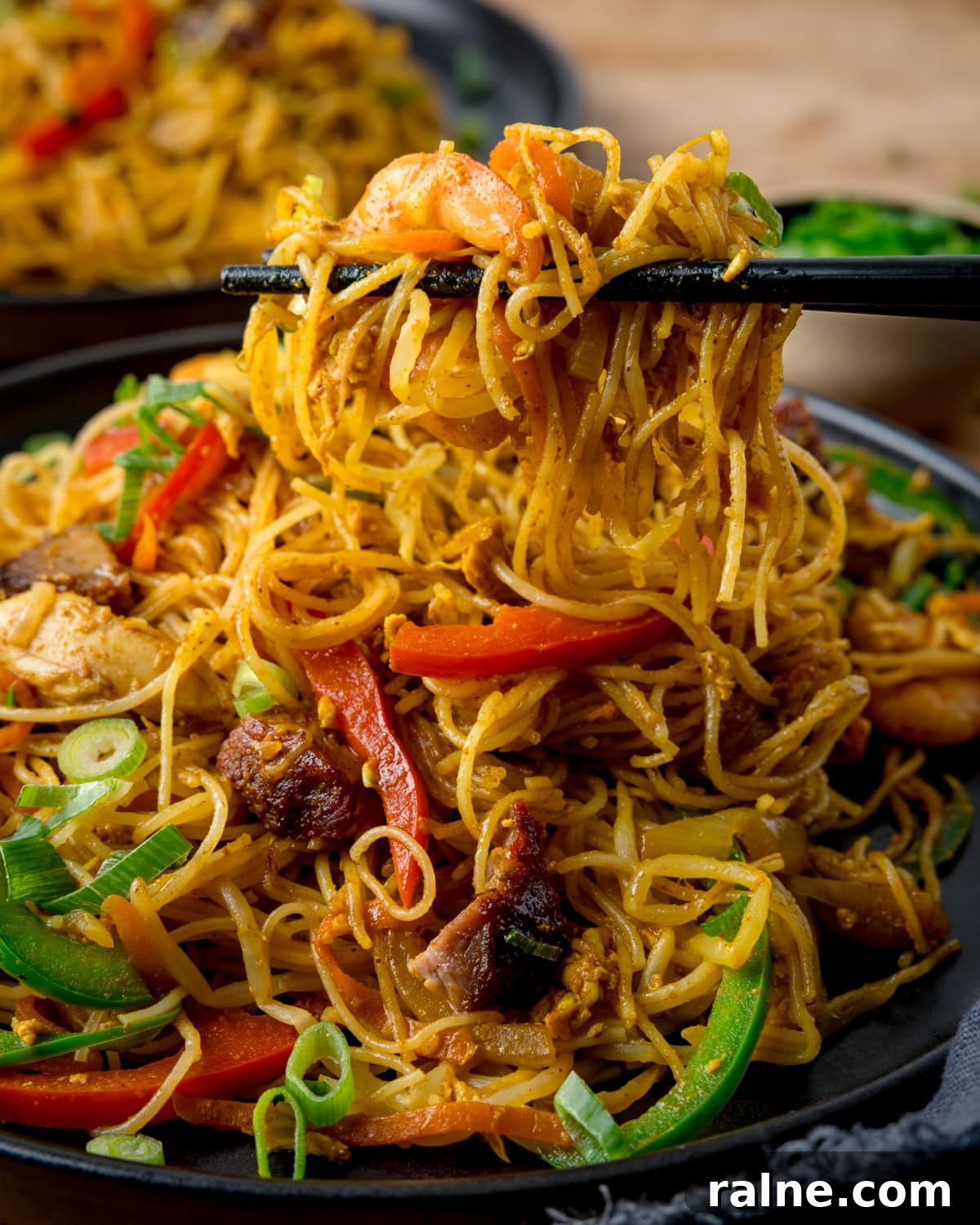 Singapore Stir-Fry Noodles with Curry and Shrimp 2 Chopsticks lifting a portion of stir-fried Singapore noodles with prawns, pork, peppers, and spring onions on a dark plate.