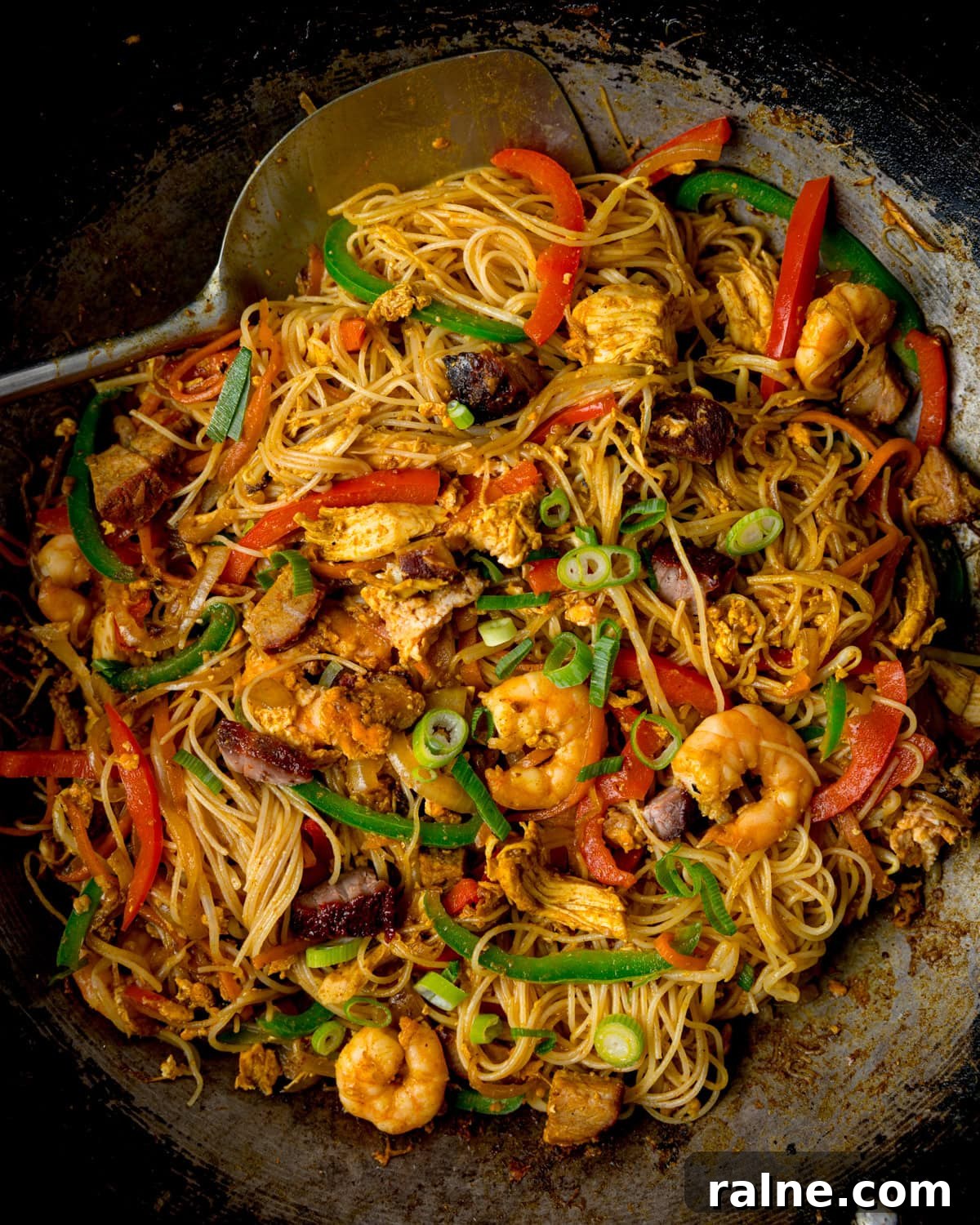 Singapore Stir-Fry Noodles with Curry and Shrimp 6 A wok filled with stir-fried rice noodles, prawns, chicken, pork, sliced vegetables, spring onions, with a large metal spatula on the side.