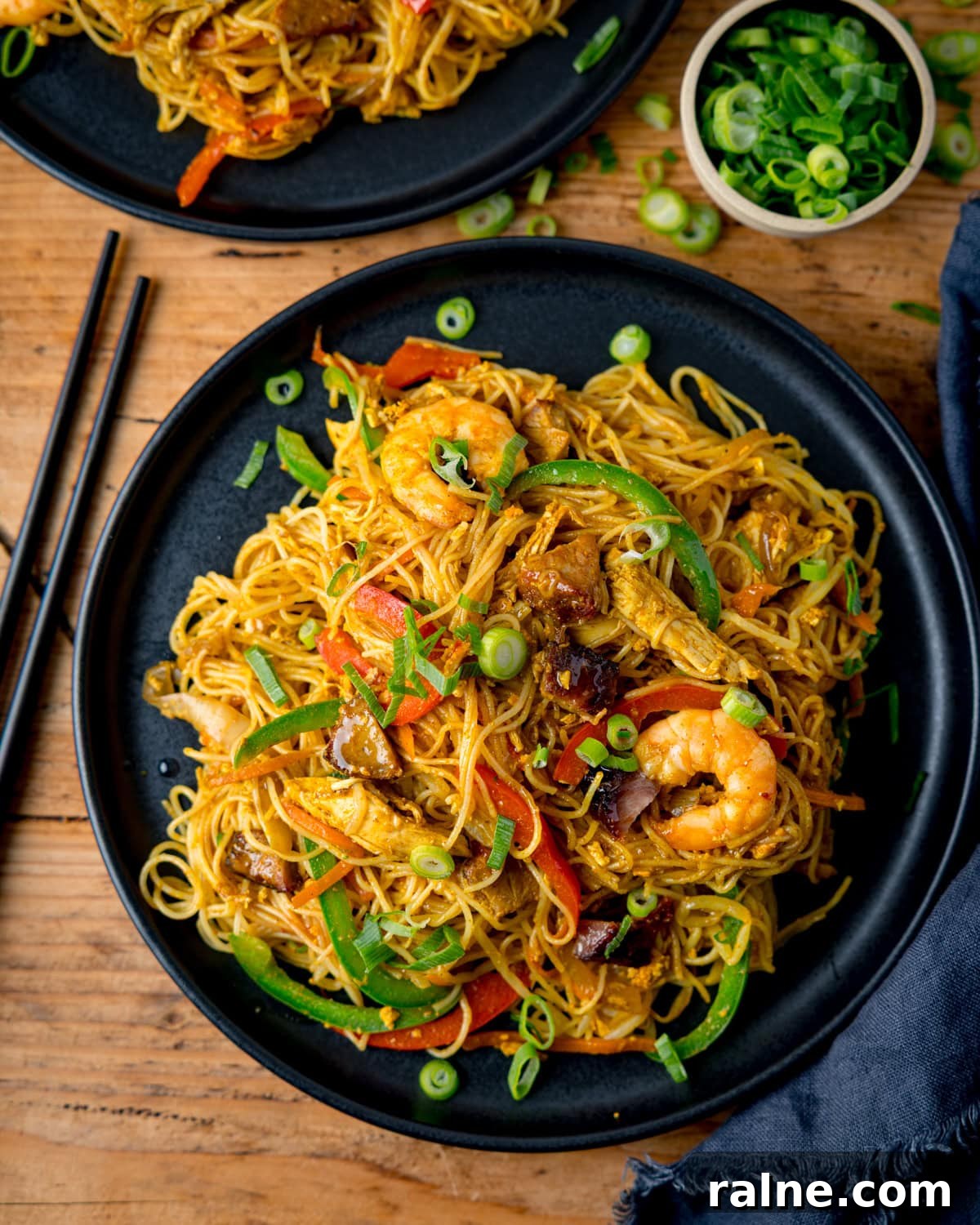 Singapore Stir-Fry Noodles with Curry and Shrimp 7 A black plate of stir-fried Singapore noodles with prawns, chicken, vegetables, and spring onions on a wooden table with chopsticks and a small bowl of sliced spring onions nearby.