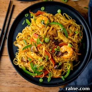 Singapore Stir-Fry Noodles with Curry and Shrimp 8 A black plate of stir-fried Singapore noodles with prawns, chicken, car siu pork vegetables, and spring onions, on a wooden table with chopsticks beside it.